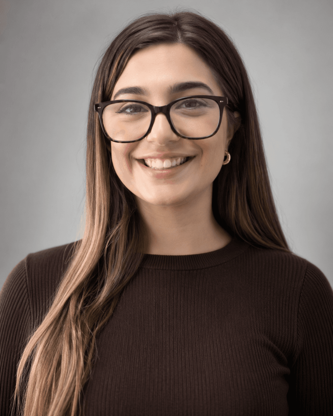 Smiling young woman with long brown hair and black glasses wearing a dark brown sweater.