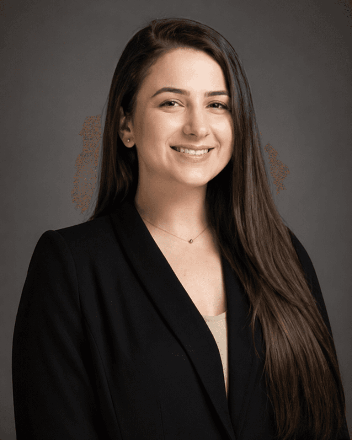 Smiling woman with long brown hair wearing a black blazer against a solid gray background.
