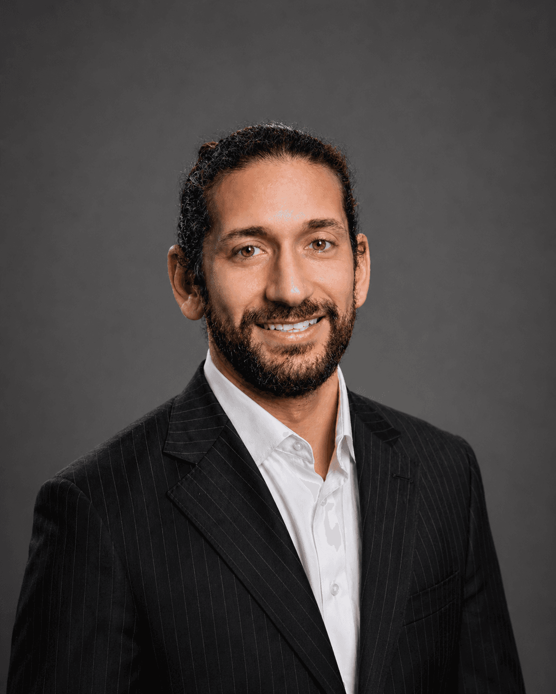 Smiling man with dark curly hair and beard wearing a pinstripe suit and white shirt.