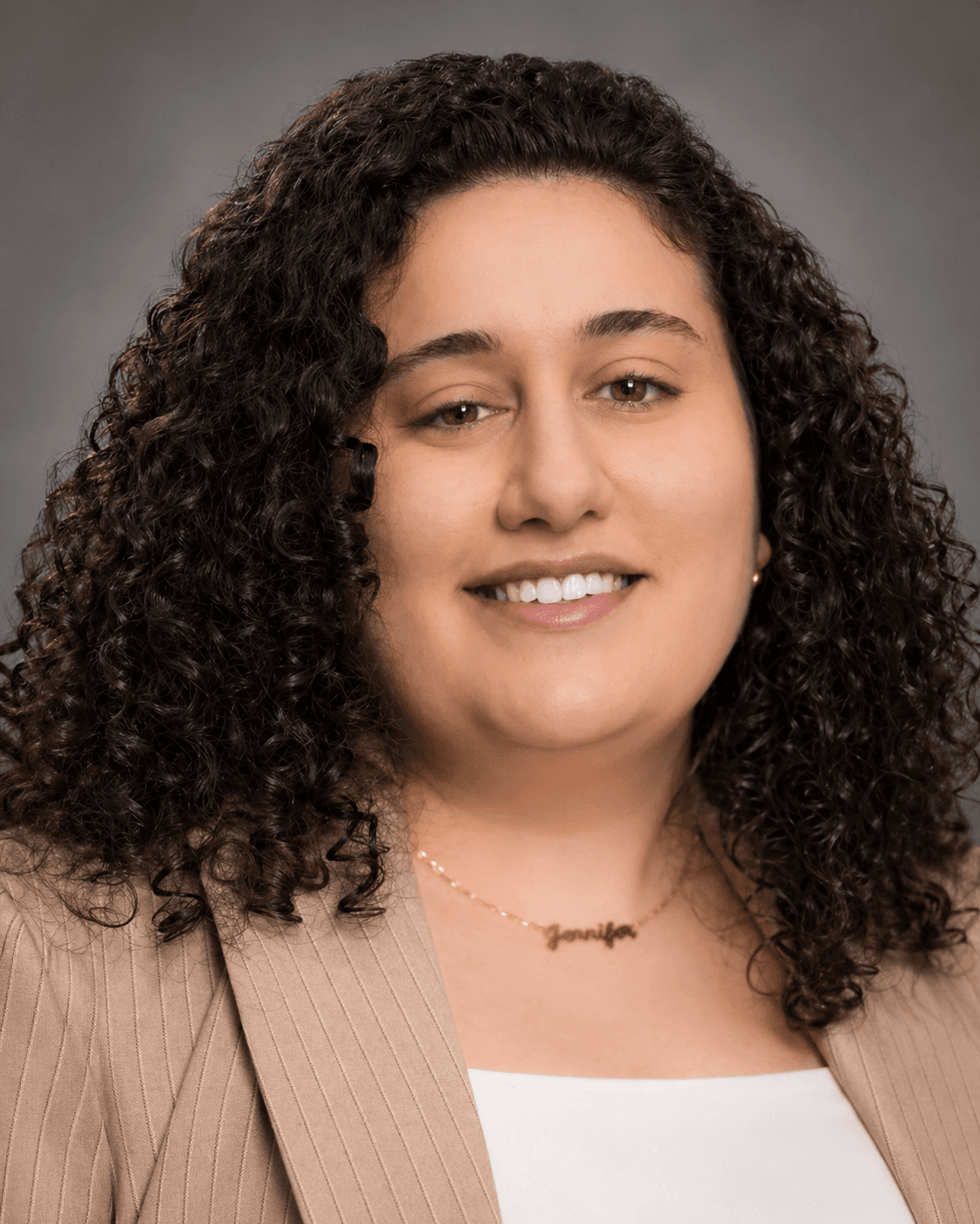 Smiling woman with dark curly hair wearing a tan pinstripe blazer and gold necklace.