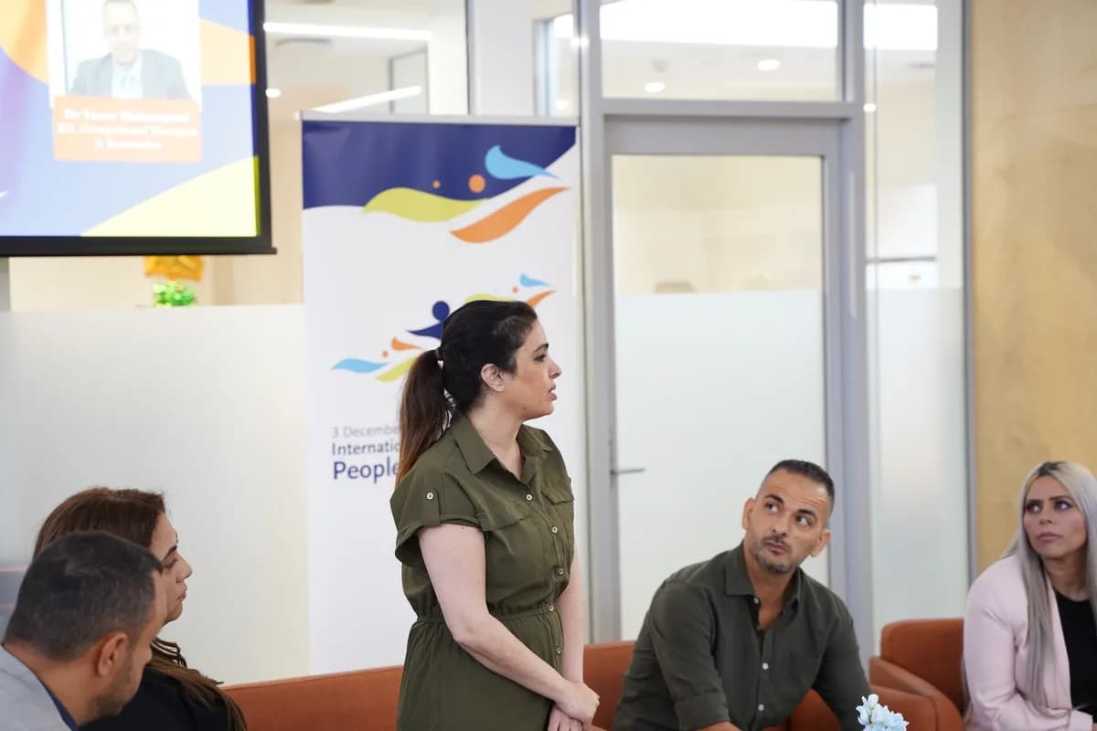 Woman in olive dress speaking to a seated audience in front of an event banner.