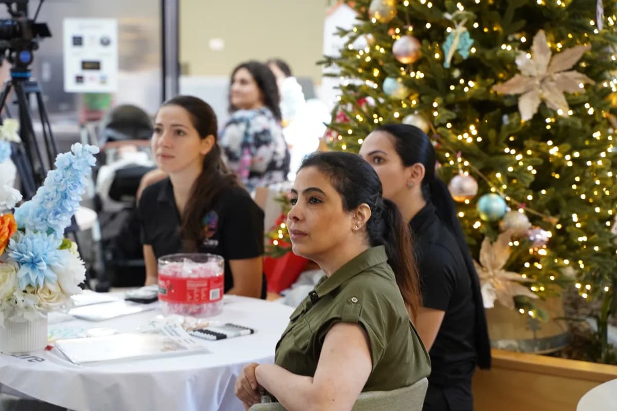 Three women sit at a table beside a decorated Christmas tree during a festive event.